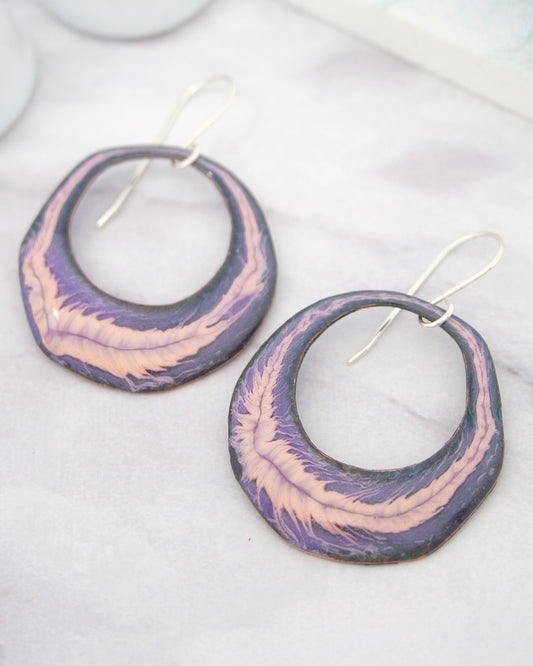 A pair of purple and pink earrings with a floral design, resting on a white surface.