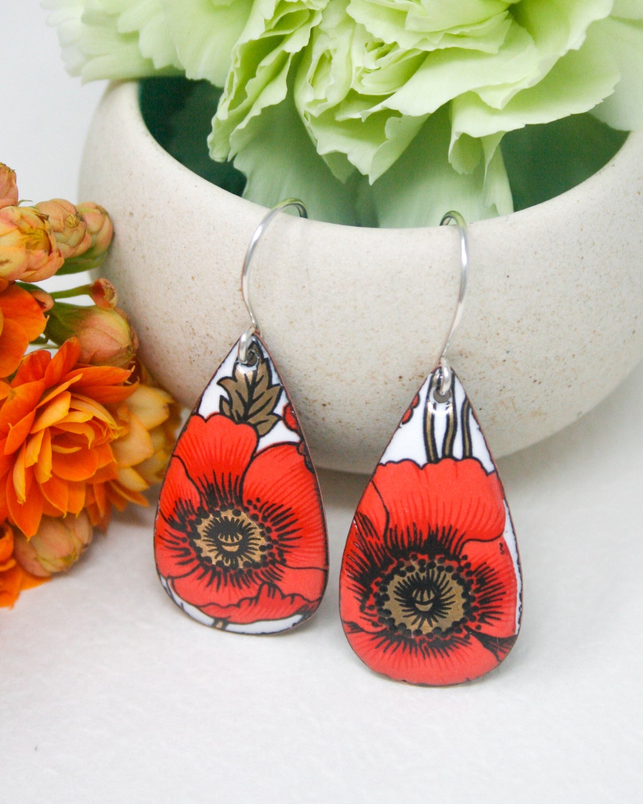 A pair of red and white earrings with floral designs, placed on a white surface next to a white ceramic container filled with green flowers.