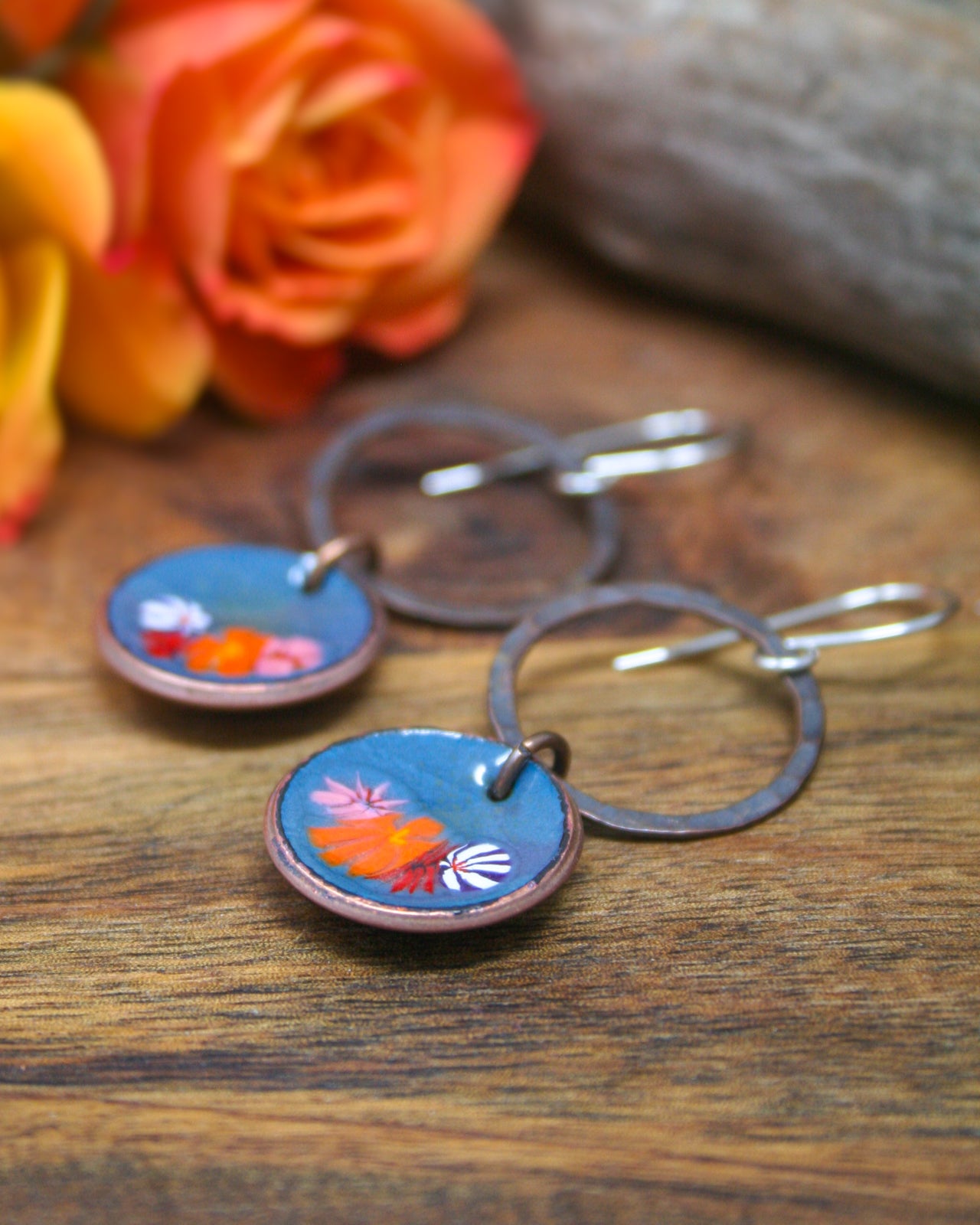 A pair of blue circular earrings with colorful floral designs, placed on a wooden surface next to a vibrant orange rose.