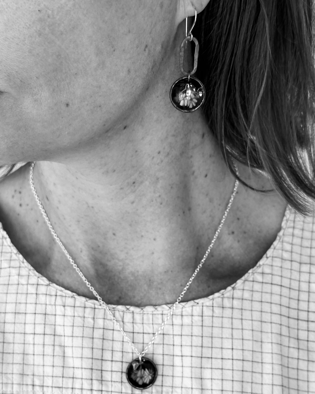 A close-up black and white image of a person's neck and chest area, featuring a necklace with a pendant and a pair of dangling earrings.