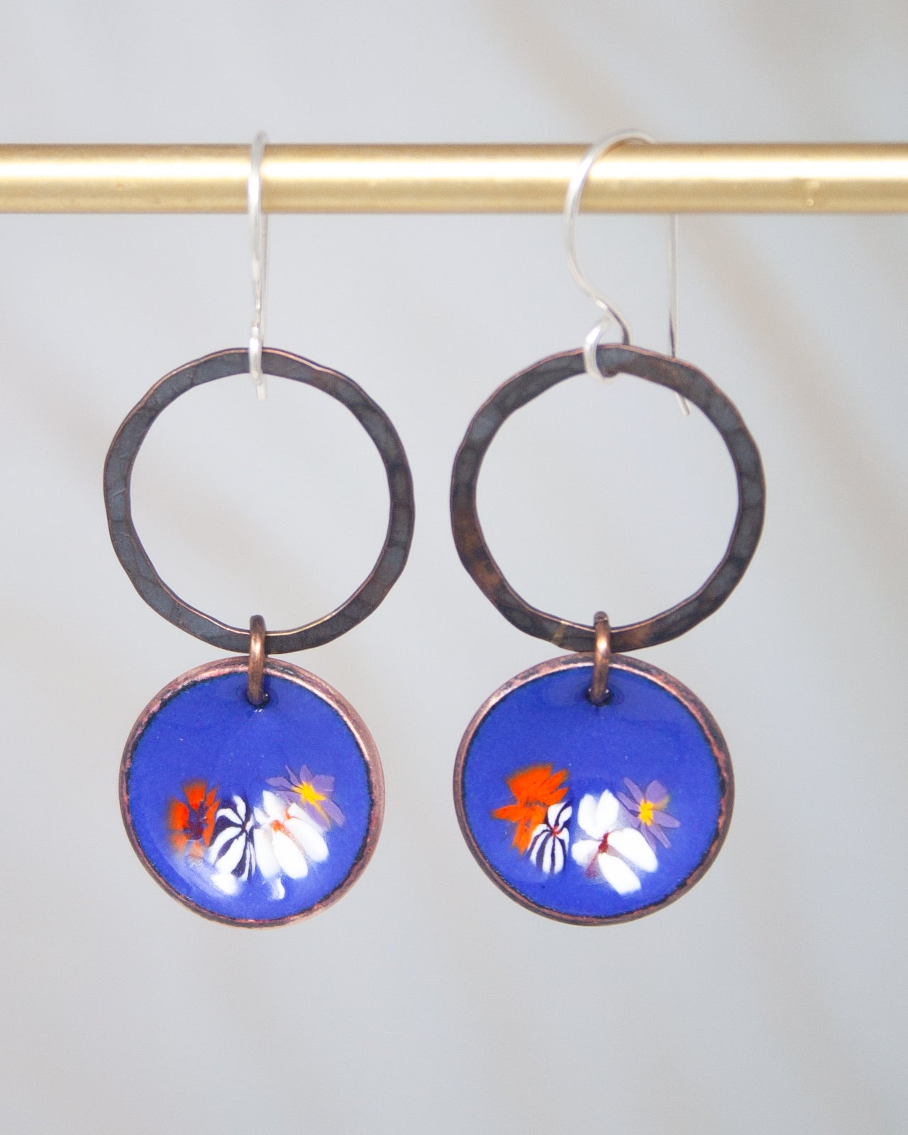 A pair of earrings with blue and orange floral designs on circular pendants, hanging from a gold-colored metal hook.