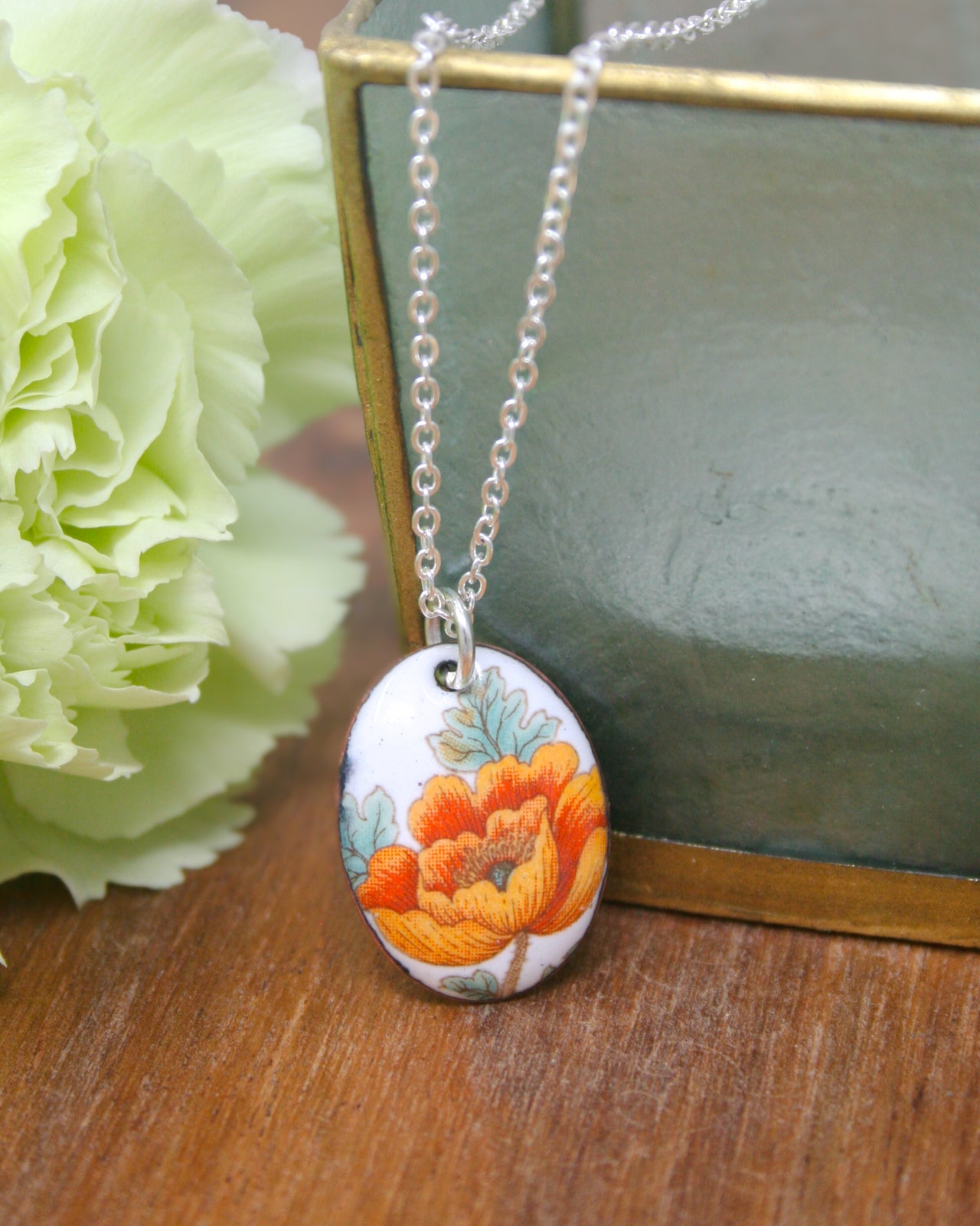 A necklace with a pendant featuring a floral design is displayed on a wooden surface, accompanied by a green vase and a white flower.