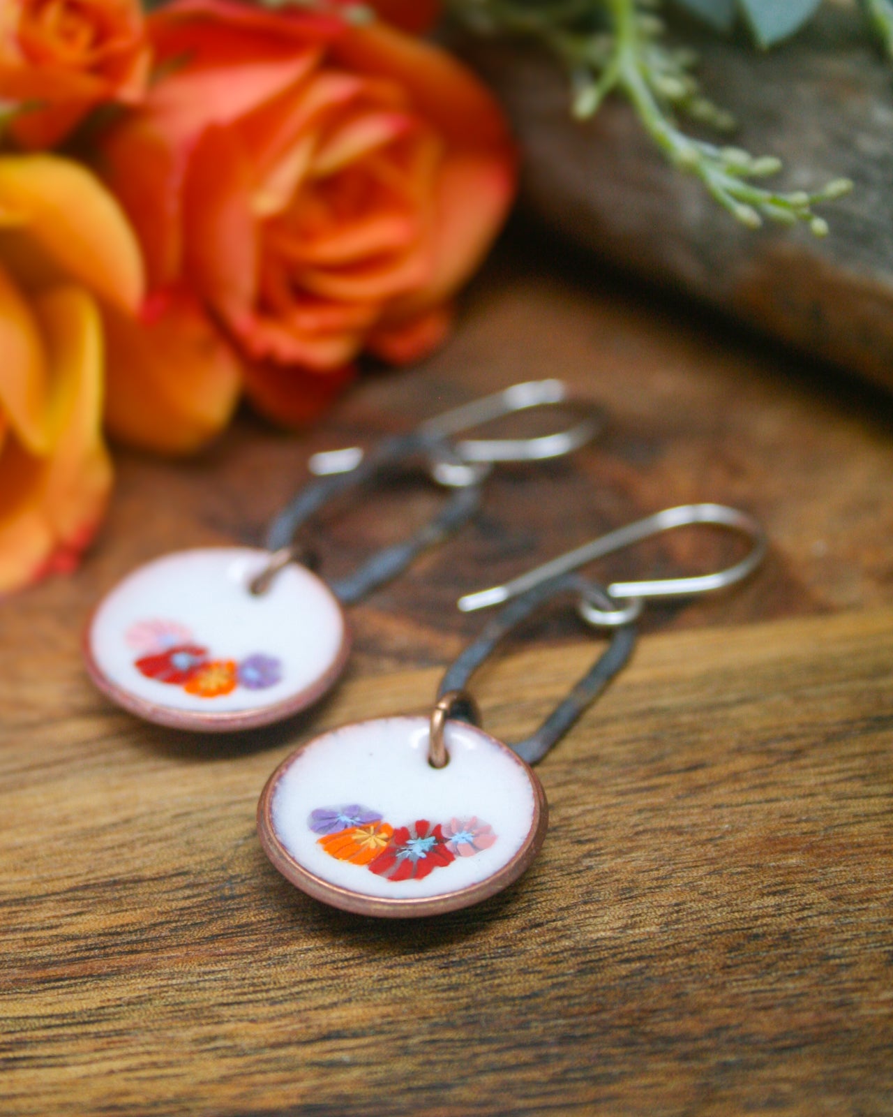 A pair of round earrings with colorful floral designs on a wooden surface, with a blurred background of orange and yellow roses.