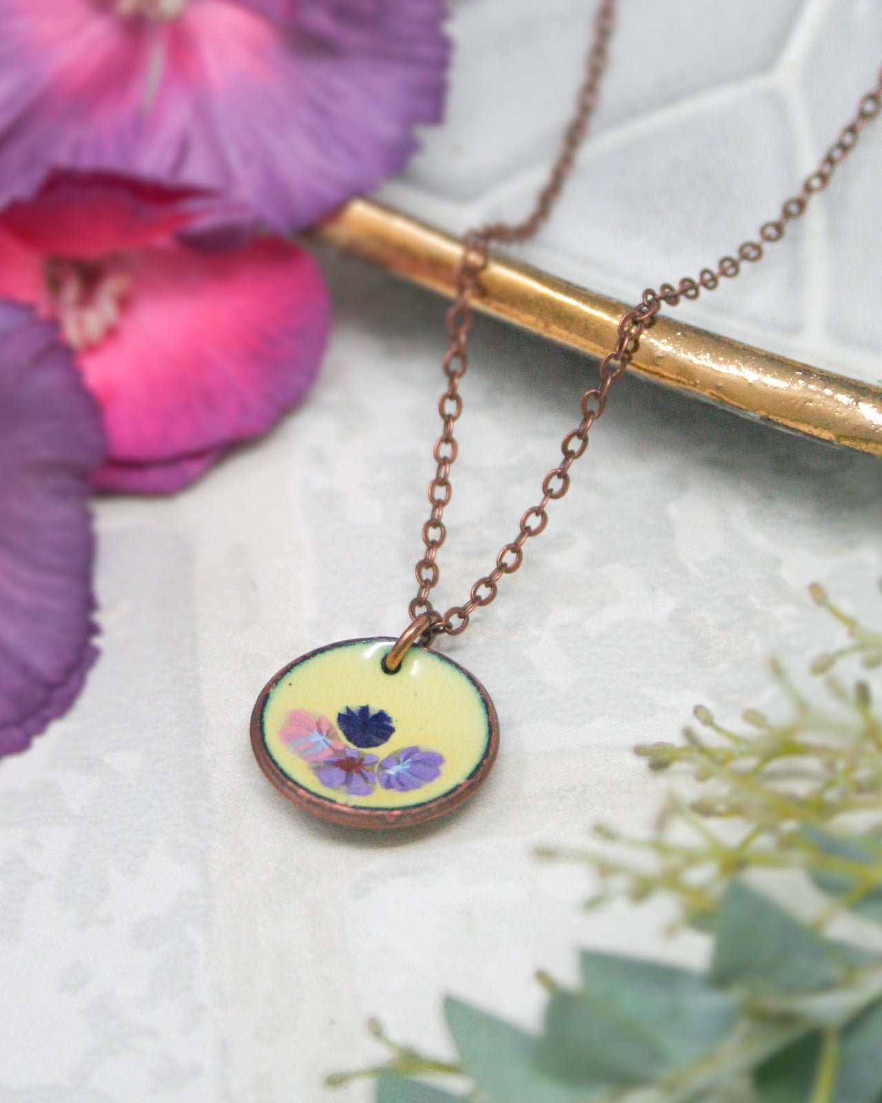 A necklace with a round pendant featuring a floral design rests on a white surface, surrounded by purple and green flowers.