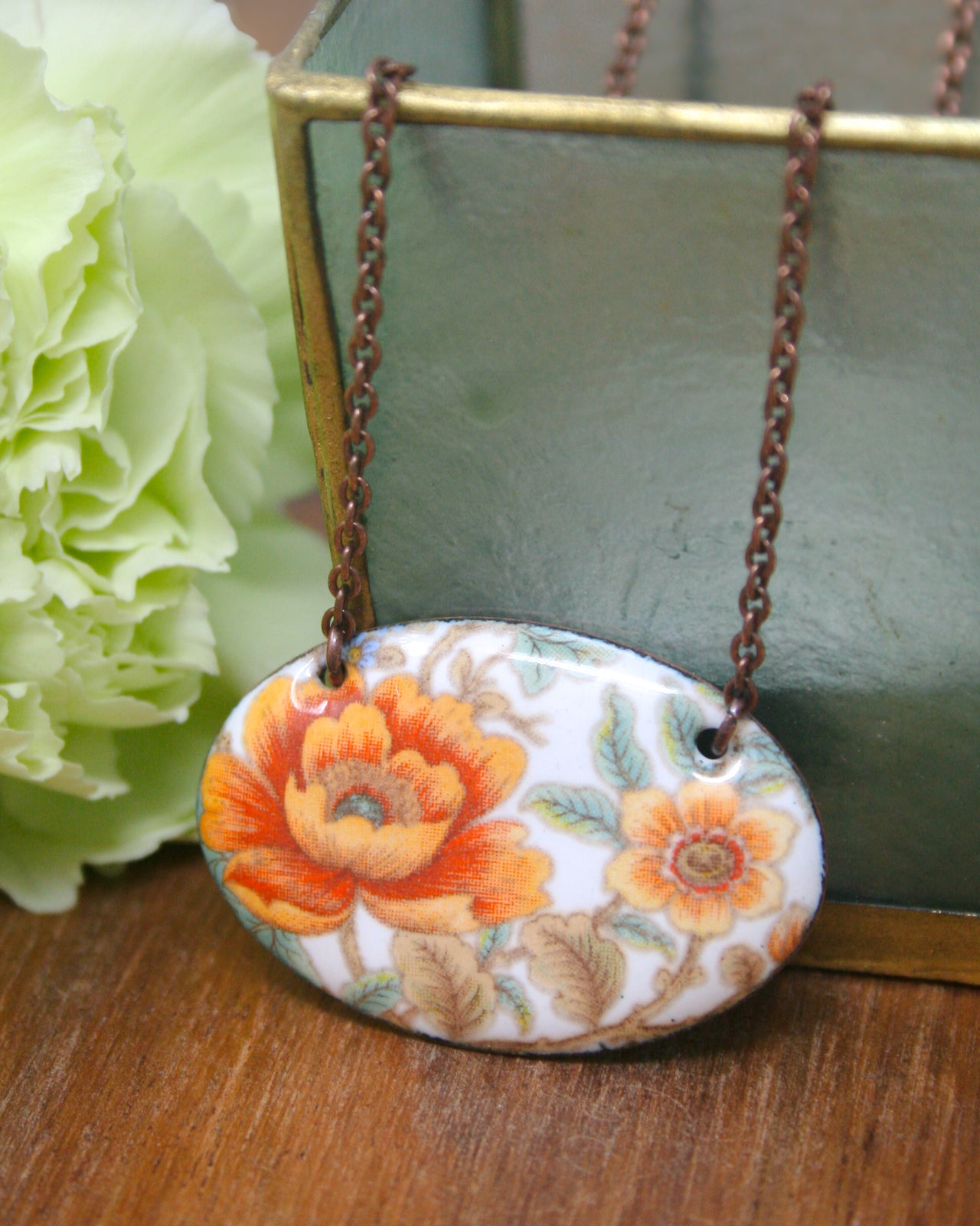 A necklace with a floral pendant is displayed on a wooden surface, accompanied by a green vase and a white flower.