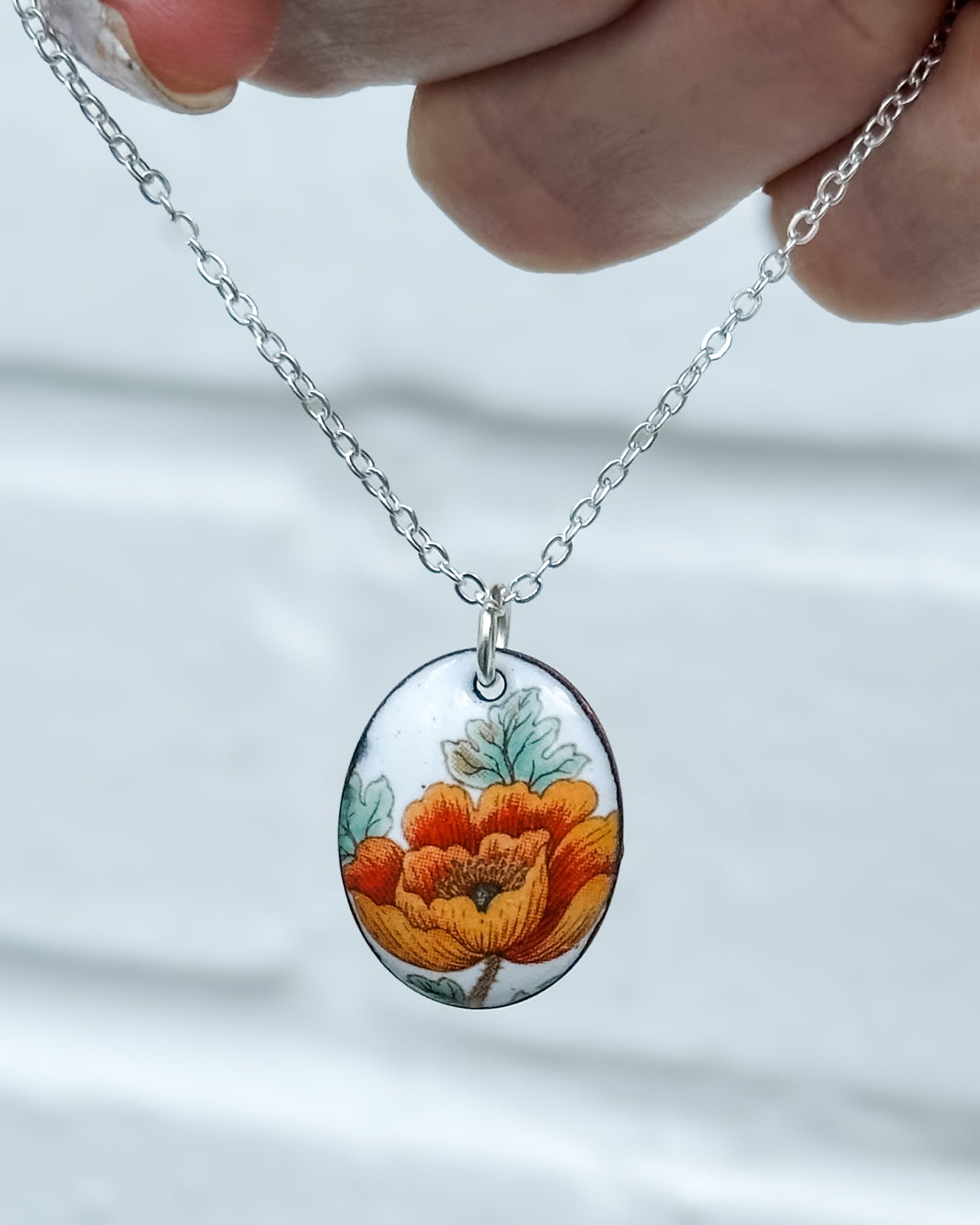 A hand is holding a silver necklace with a pendant featuring a colorful floral design.
