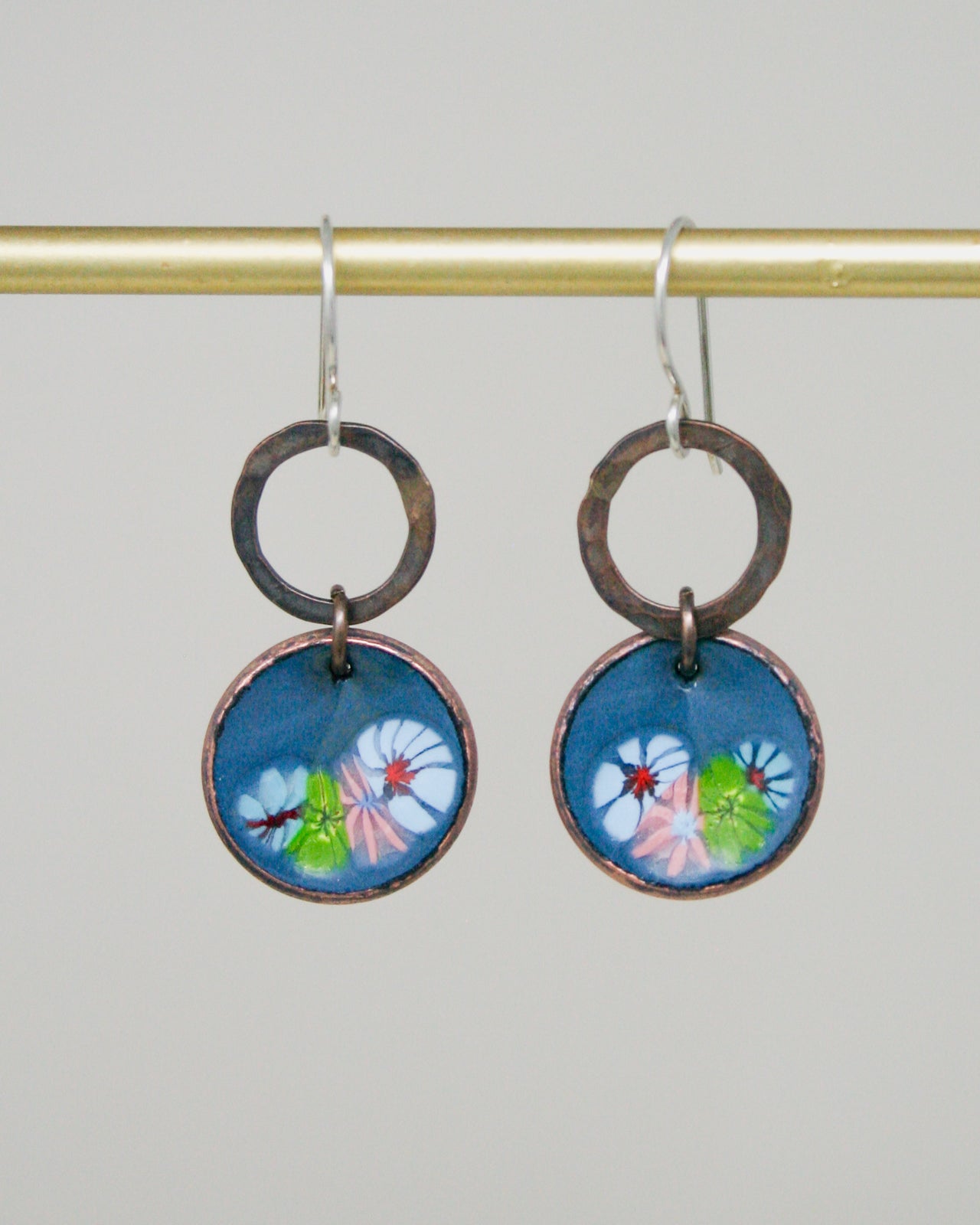 A pair of earrings with a blue circular design featuring colorful flowers hanging from a gold-colored metal hook.