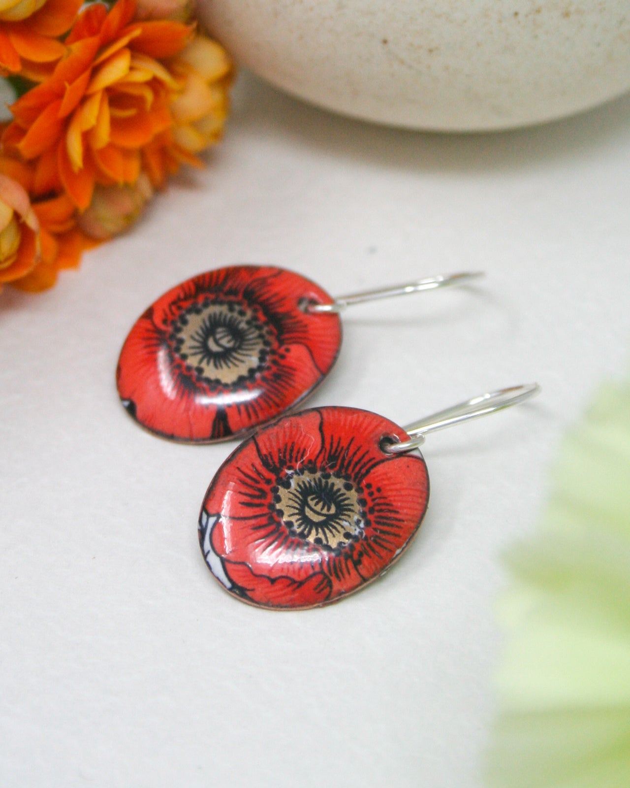 A pair of red earrings with a floral design, placed on a white surface next to a white ceramic object and some orange flowers.