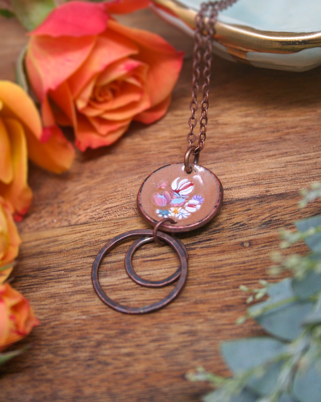 a necklace with a circular pendant, adorned with a floral design, resting on a wooden surface. The necklace is surrounded by a variety of flowers, including a large orange rose and smaller orange flowers, creating a vibrant and colorful scene.