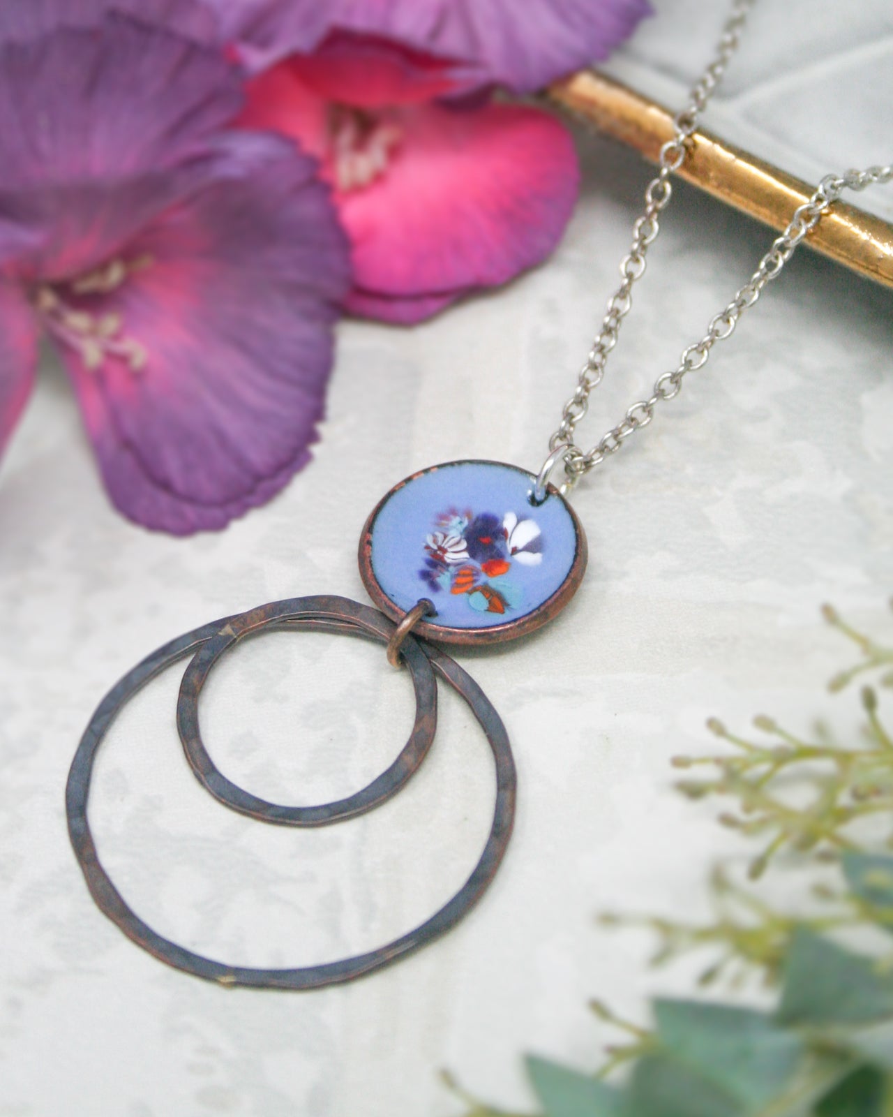 a necklace with a circular pendant, which appears to be a piece of jewelry. The pendant is surrounded by two large circular rings, creating a unique and eye-catching design. The necklace is placed on a white surface, with a few purple flowers visible in the background.