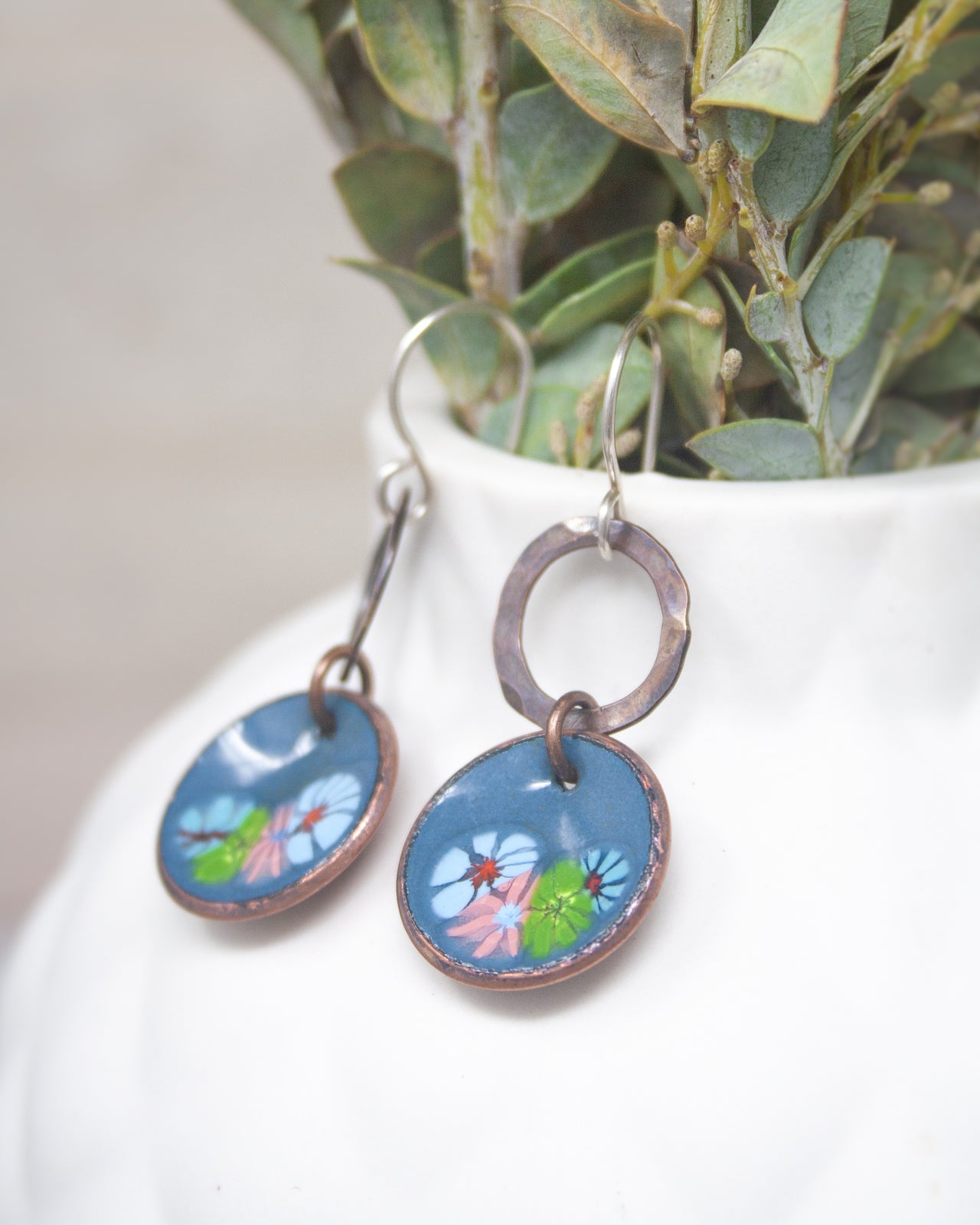 A pair of blue earrings with floral designs hangs from a hook, placed in a white vase with green leaves.