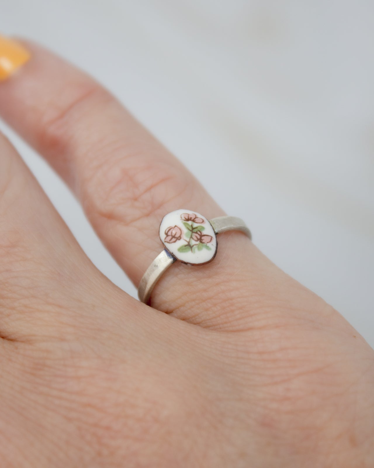 A close-up of a person's hand wearing a silver ring with a small oval-shaped charm featuring a floral design.