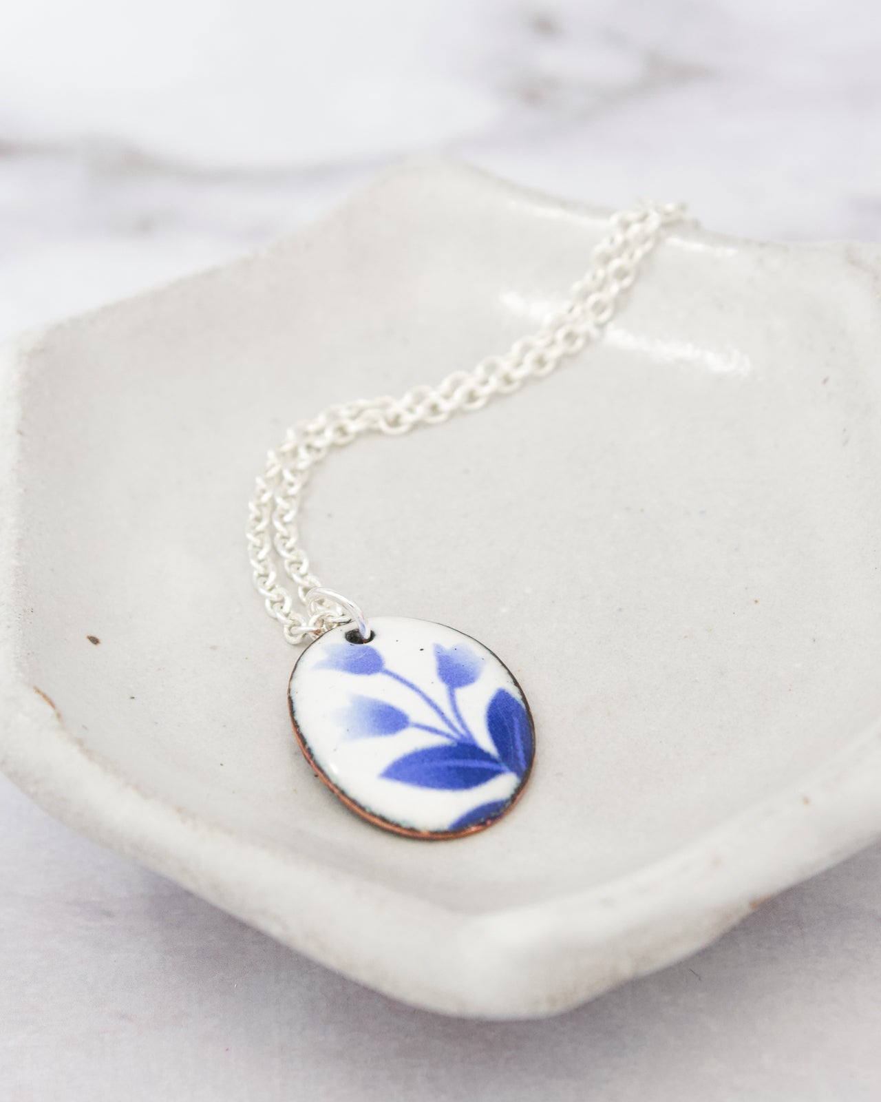 A white ceramic dish containing a silver necklace with a blue floral pendant.