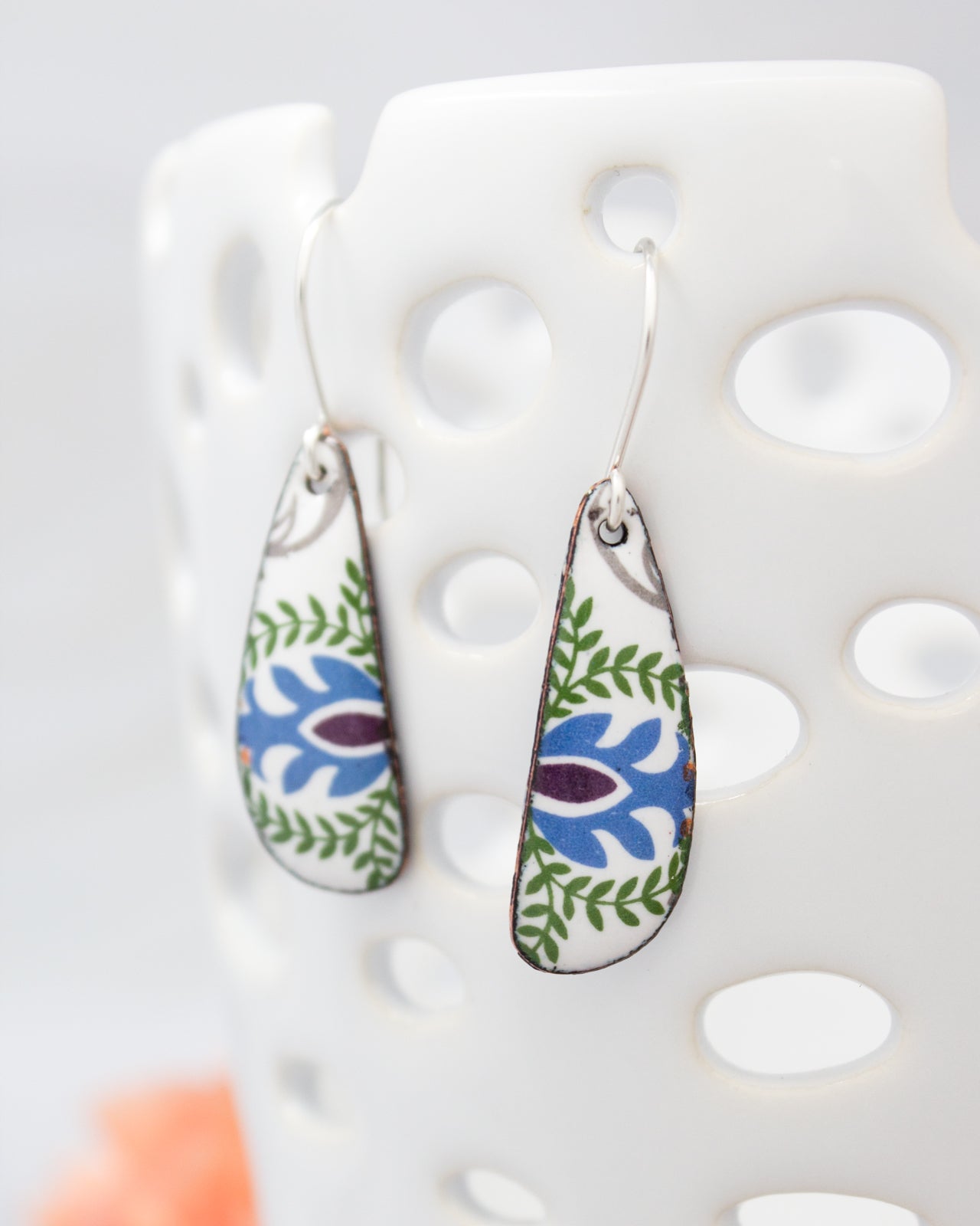 A pair of earrings with a floral design on the backings, hanging from a white perforated stand.