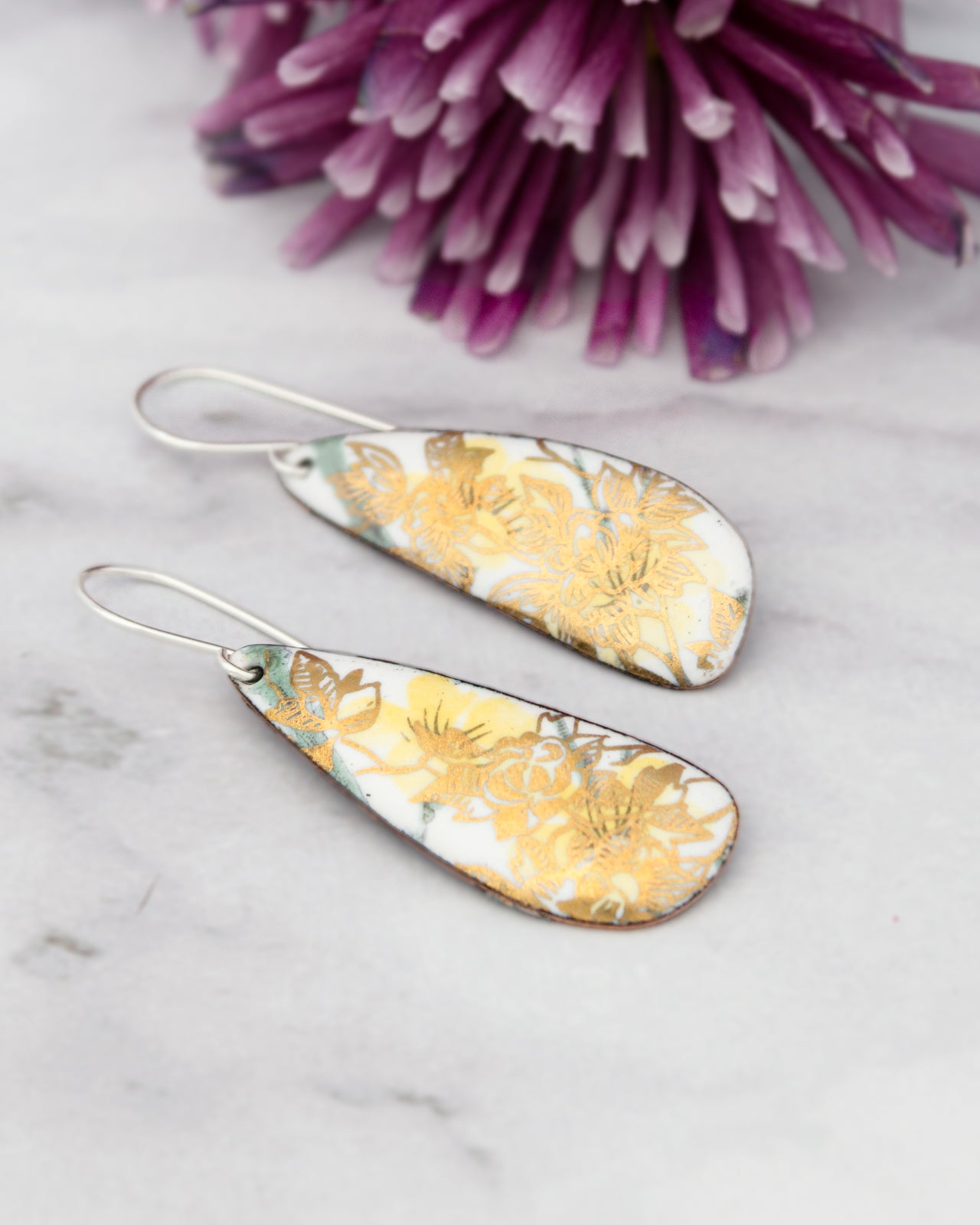a pair of earrings with a floral design, placed on a marble surface next to a purple flower.