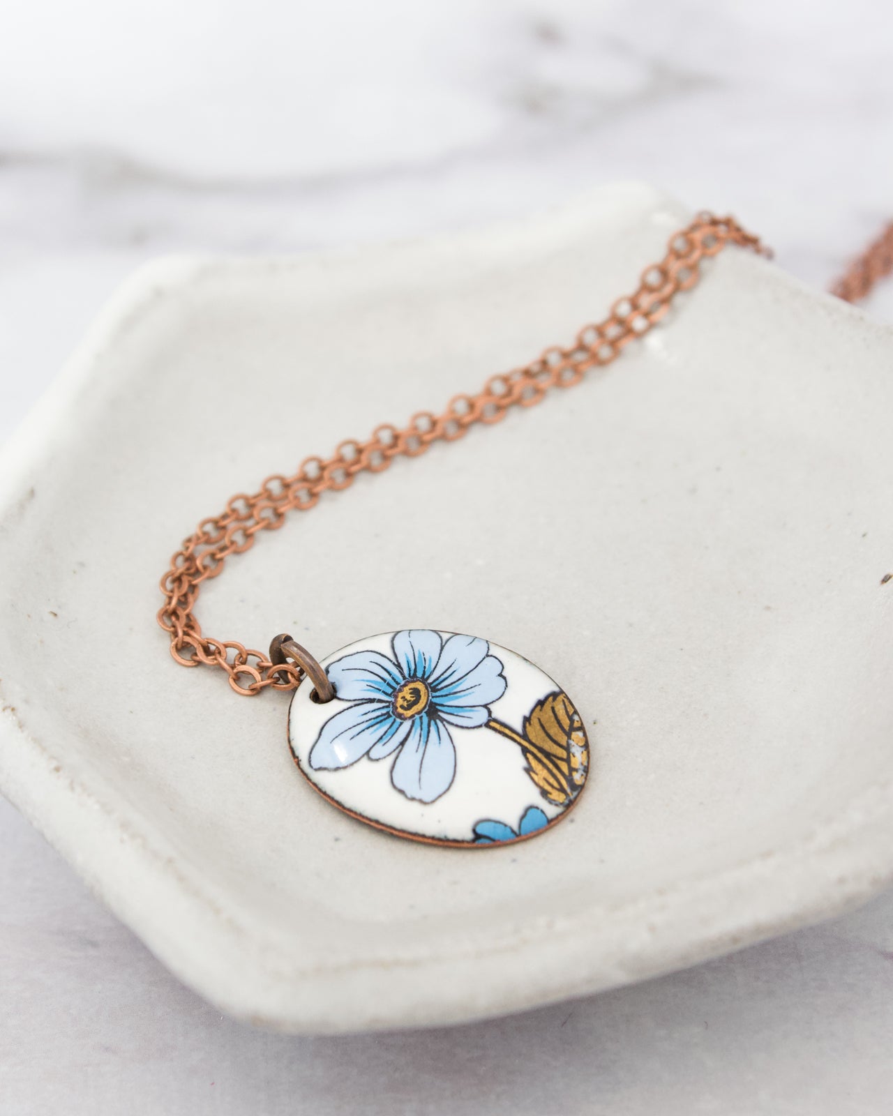 A necklace with a pendant featuring a blue flower design is displayed on a white plate.