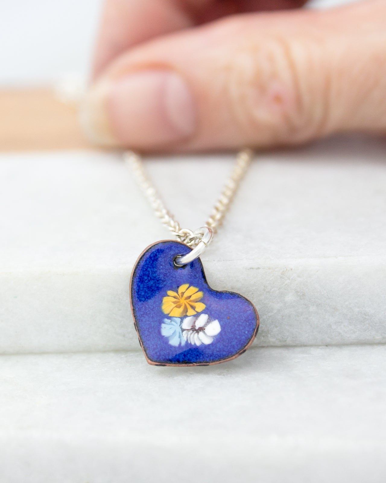 A hand is holding a heart-shaped pendant with a blue background and yellow flowers.