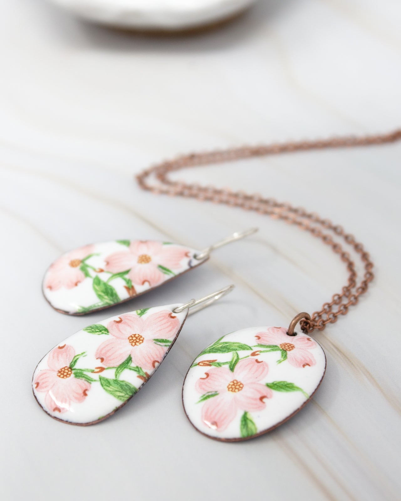 a pair of earrings and a necklace with floral designs, including pink flowers and green leaves, displayed on a marble surface.