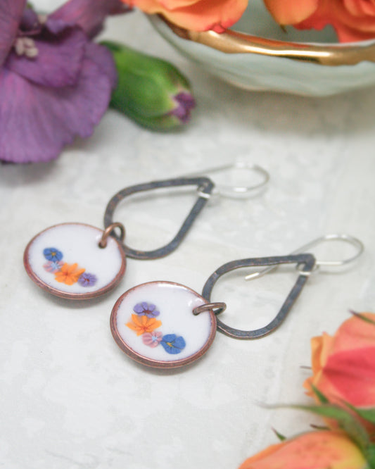 A pair of earrings with floral designs on the circular pendants, placed on a white surface alongside a bowl of orange flowers.