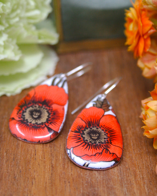 a pair of earrings with a floral design, including red and orange flowers, placed on a wooden surface.