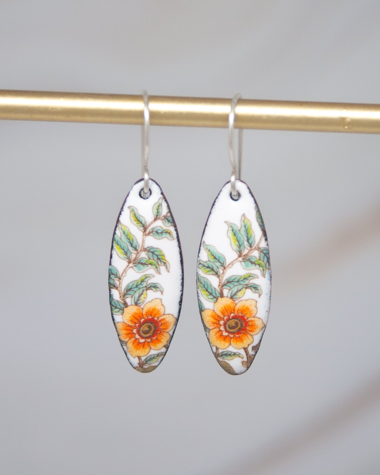 A pair of earrings with a floral design, featuring a white background with a vibrant orange and green flower and leaves pattern.