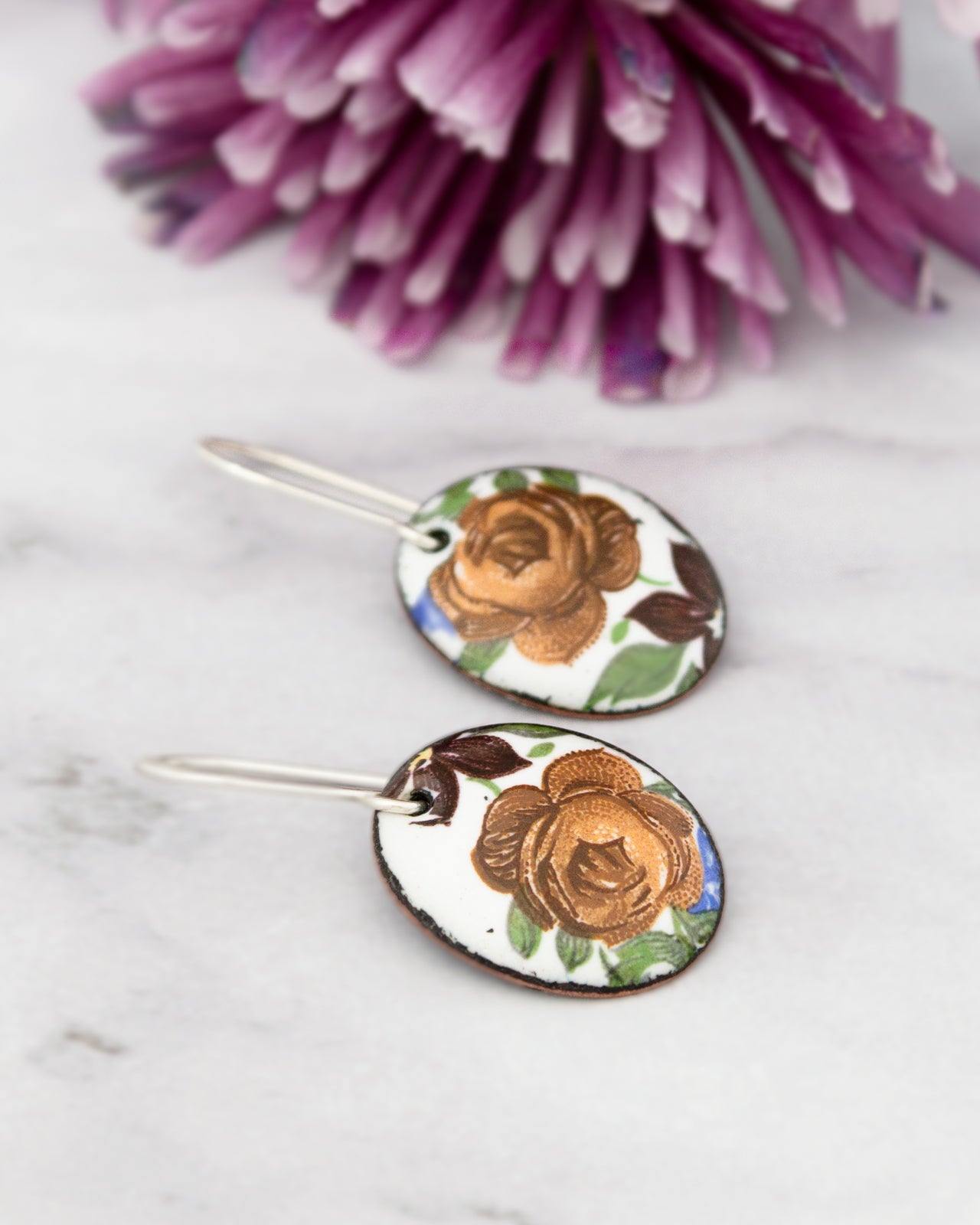 two oval-shaped earrings with a floral design, placed on a white marble surface.
