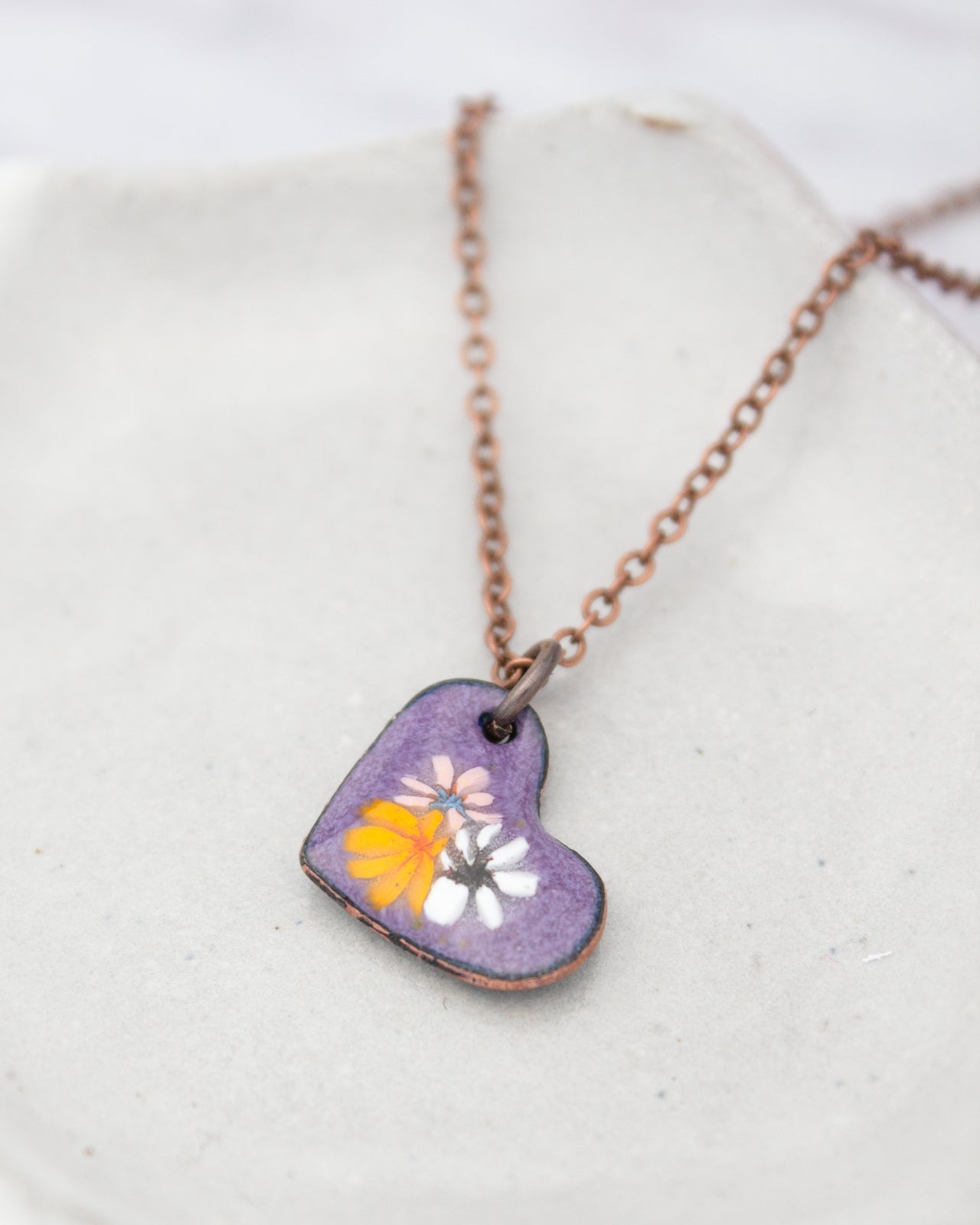A heart-shaped pendant necklace with a purple background and colorful flowers is displayed on a white surface.