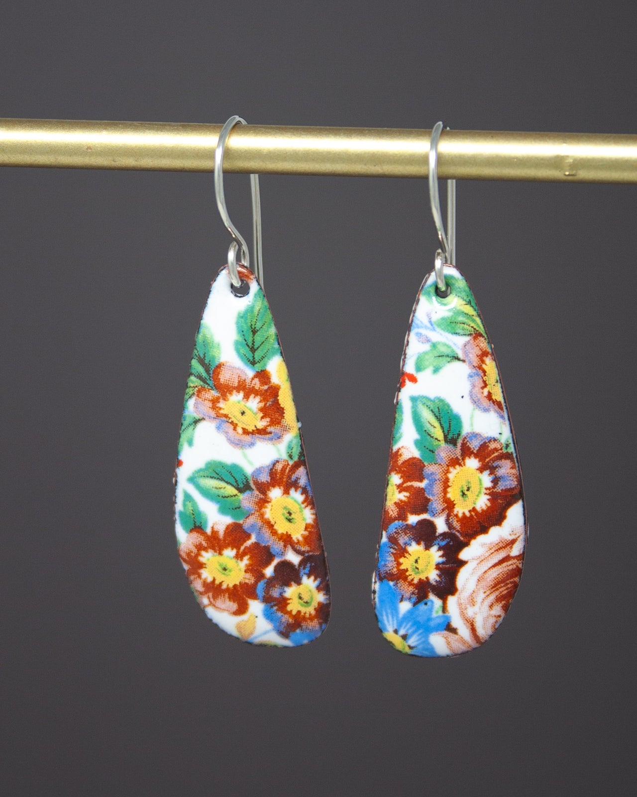 A pair of colorful earrings with floral designs hanging from a gold-colored metal hook.