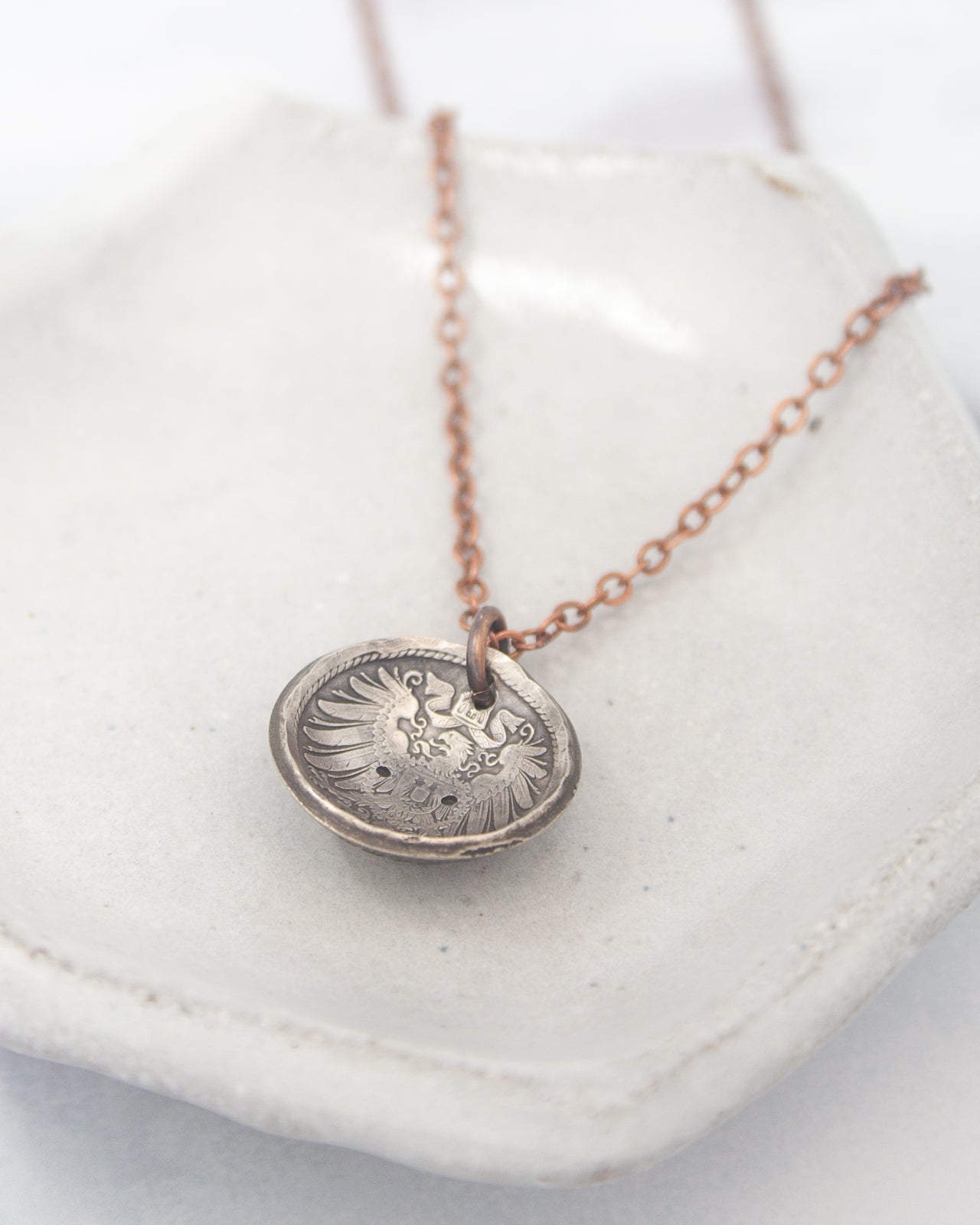 A silver-colored pendant necklace with a circular pendant featuring an intricate design, resting on a white surface.