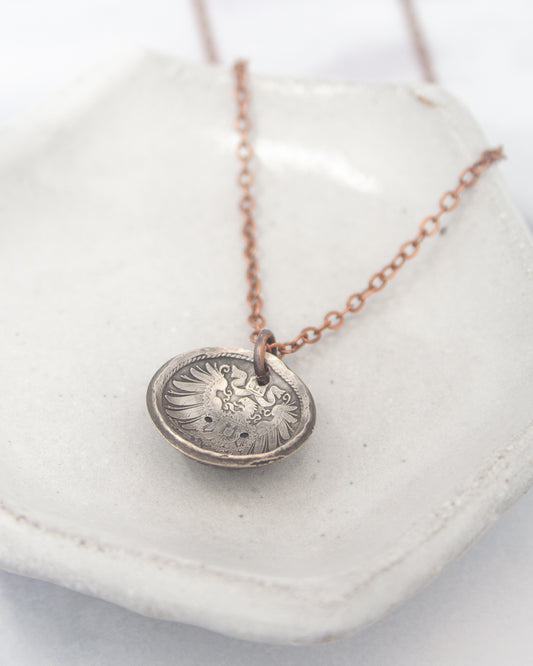 A silver-colored pendant necklace with a circular pendant featuring an intricate design, resting on a white surface.