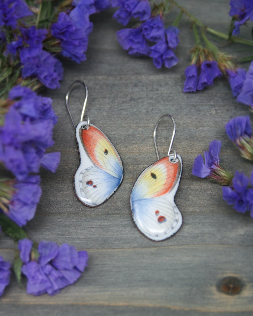 A pair of earrings with butterfly wing designs, placed on a wooden surface surrounded by purple flowers.