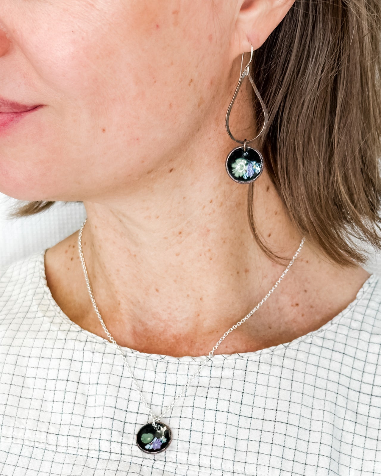 A woman wearing a necklace with a pendant and earrings, and a white shirt with a patterned design.