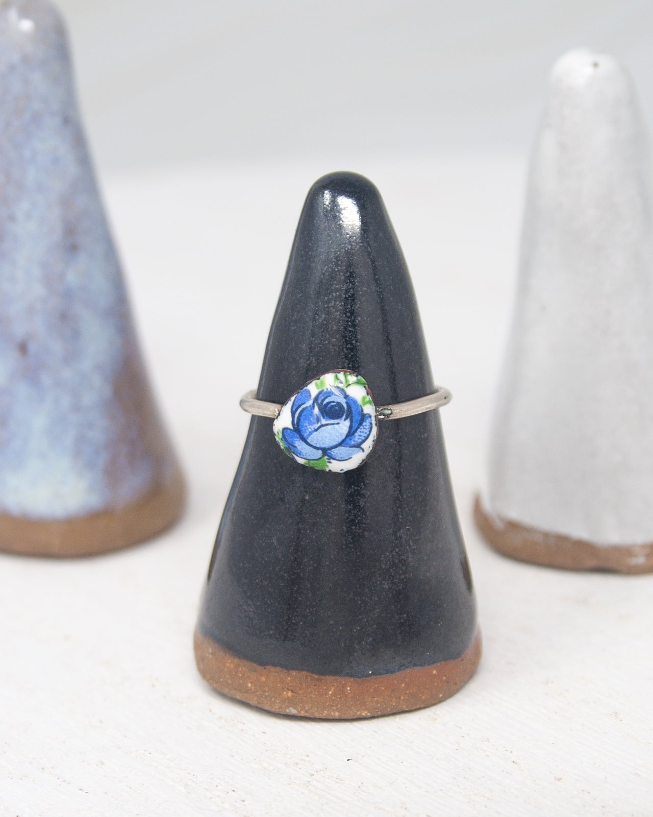 A black ceramic ring stand with a silver ring featuring a blue flower design on it, placed between two white ceramic objects.