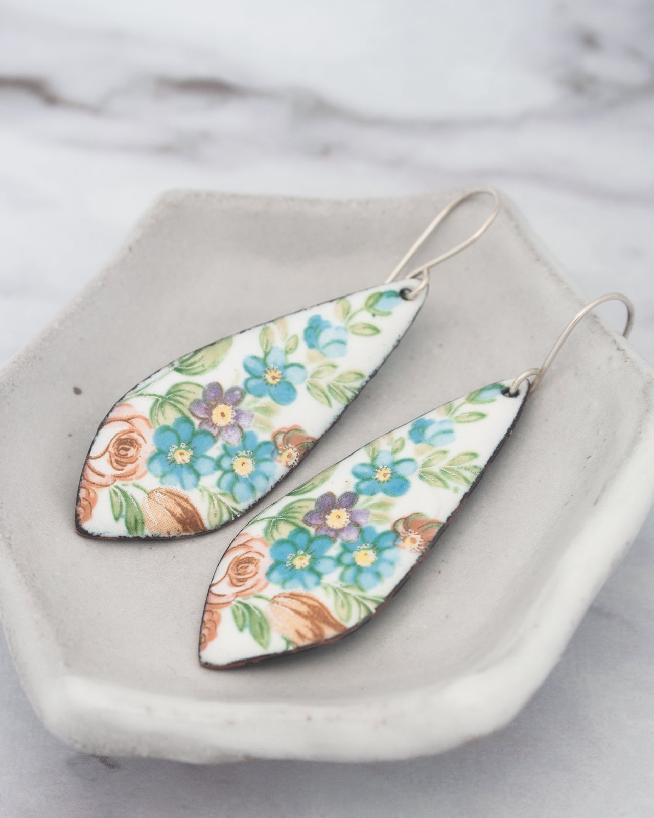 A pair of earrings with a floral design, featuring blue, orange, and white flowers, are displayed on a white plate.
