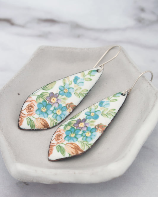 A pair of earrings with a floral design, featuring blue, orange, and white flowers, are displayed on a white plate.