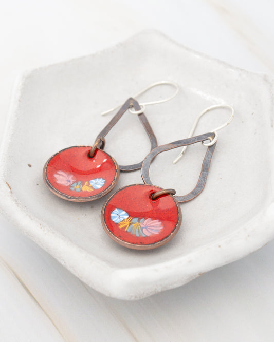 A pair of red earrings with floral designs on the circular pendants, displayed on a white plate.