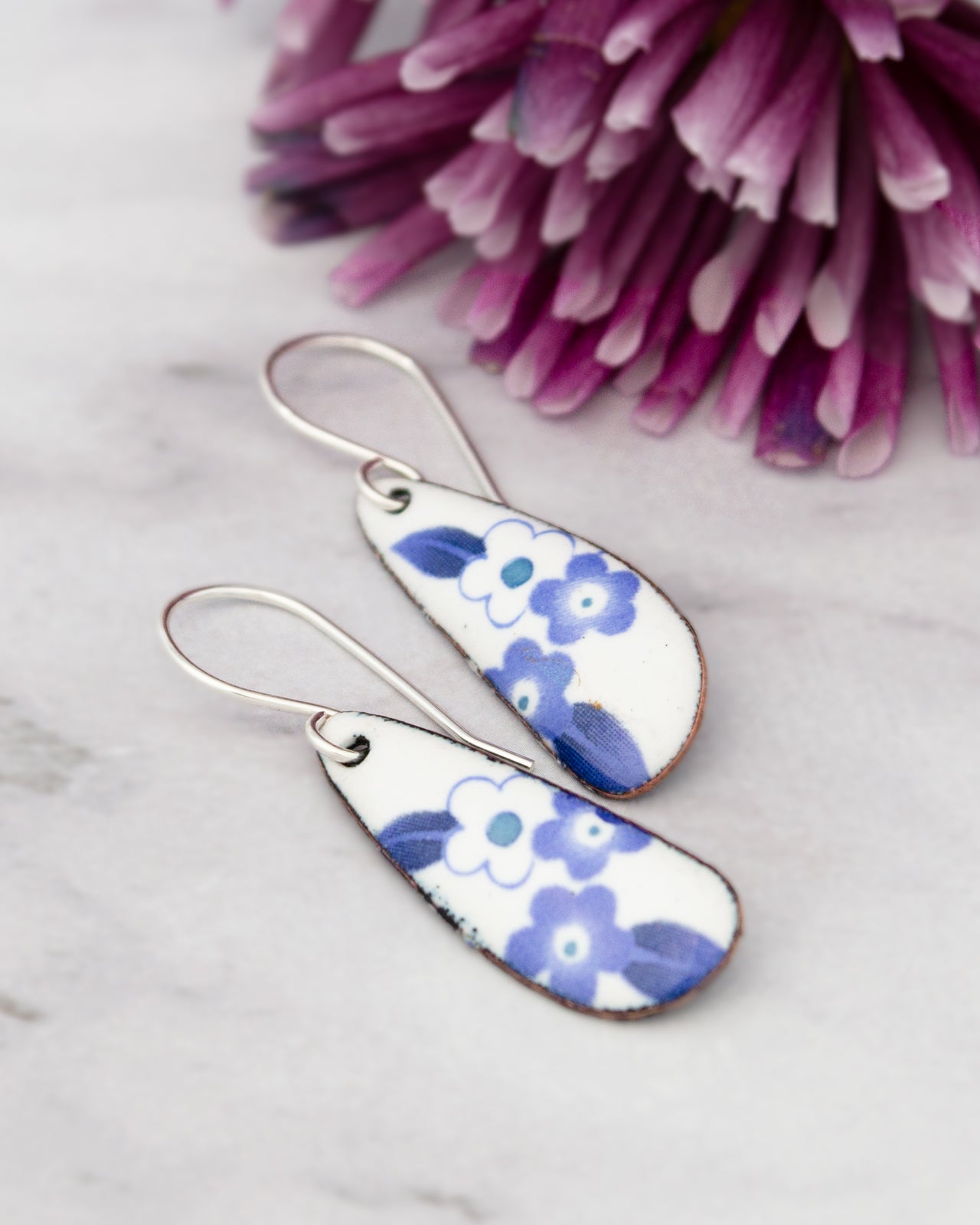 A pair of earrings with blue floral designs rests on a marble surface, accompanied by a cluster of purple flowers in the background.