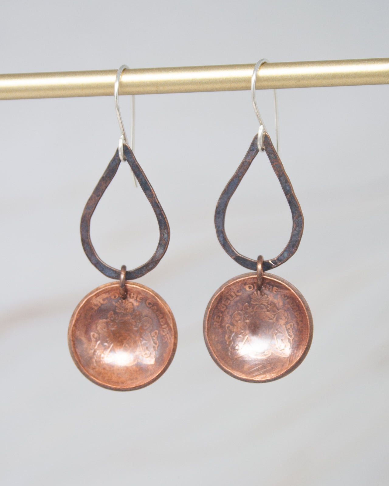 A pair of copper-colored earrings with a teardrop-shaped design and a round metal pendant hanging from a gold-colored metal hook.