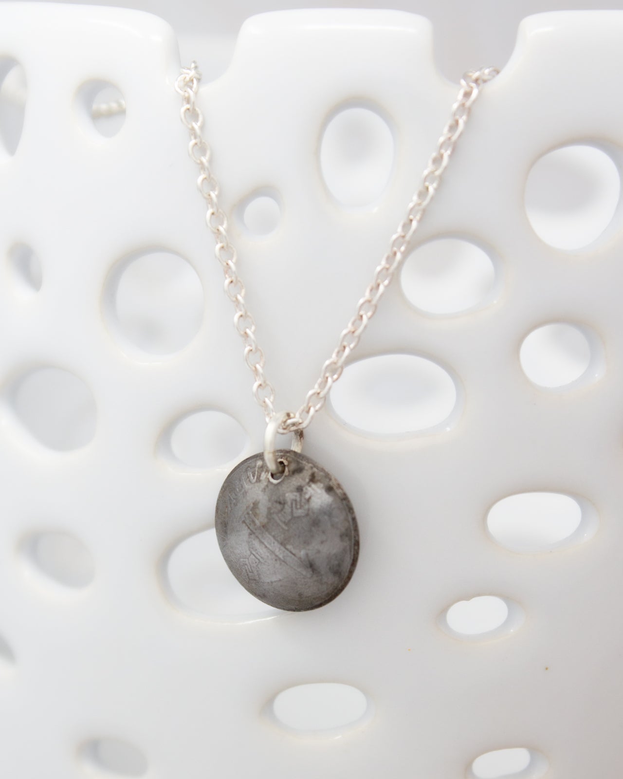A silver necklace with a round pendant is displayed against a white background with a perforated pattern.