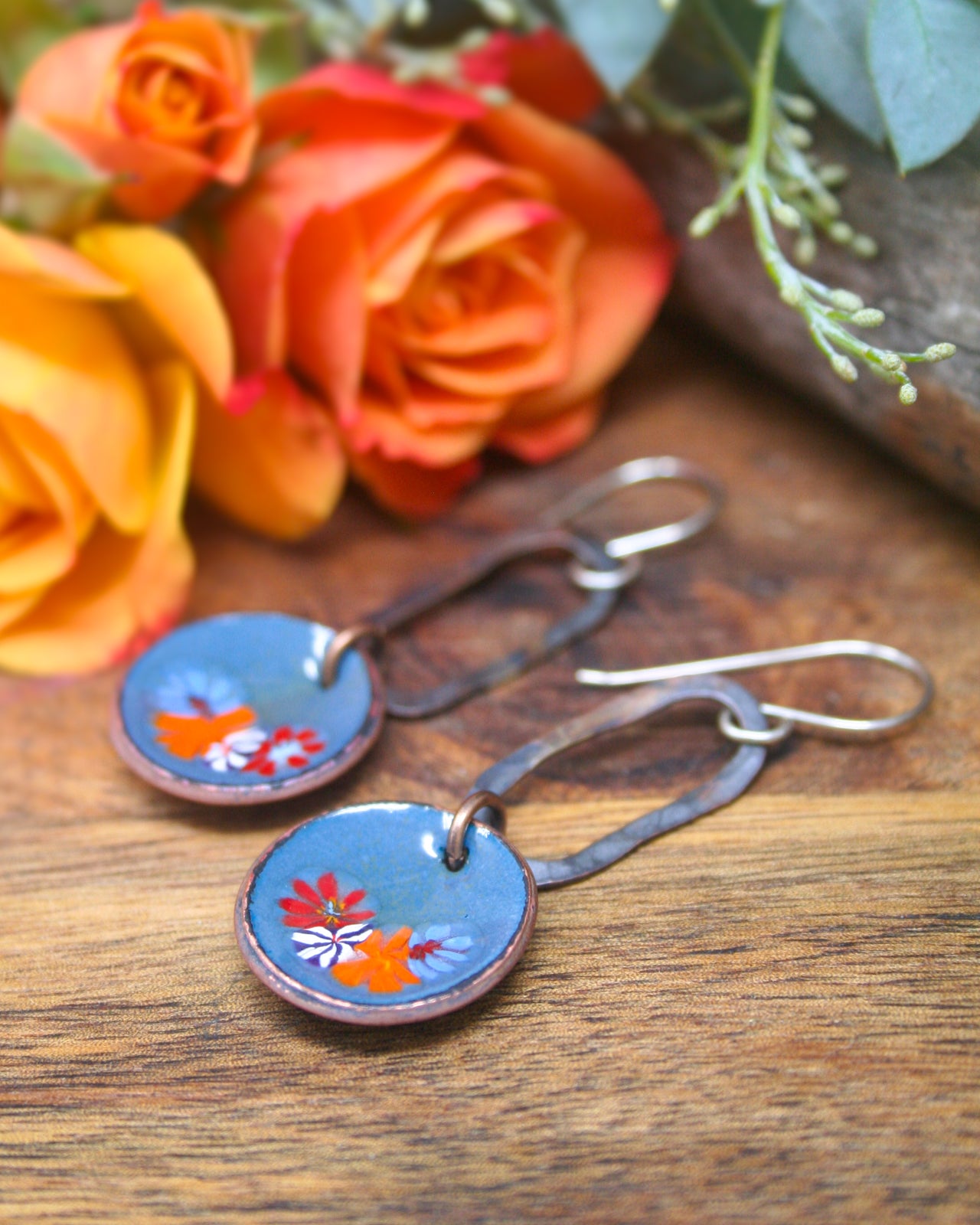 A pair of blue earrings with floral designs rests on a wooden surface, accompanied by a bouquet of orange roses and greenery.