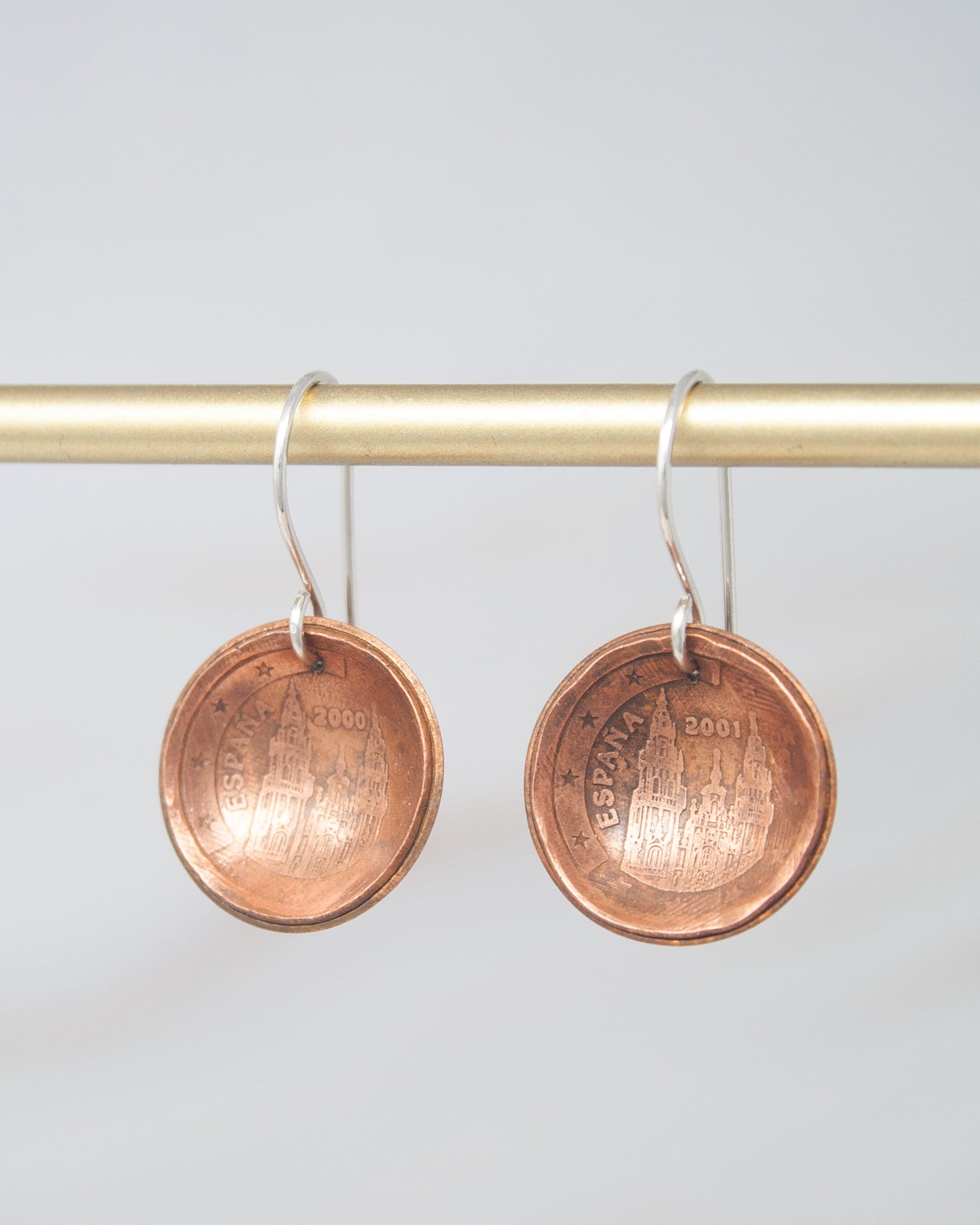 A pair of copper-colored earrings with a design on the surface, hanging from a gold-colored metal hook.