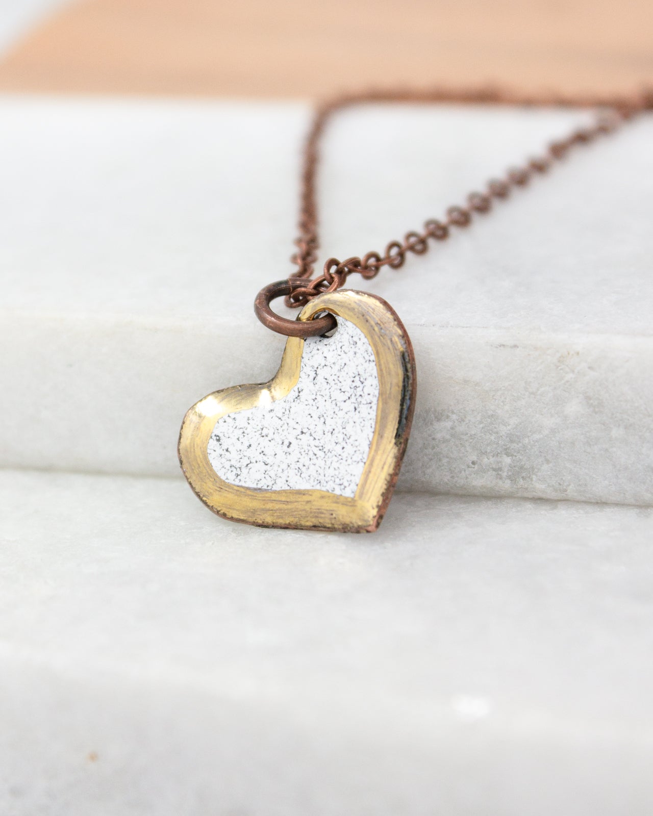 A heart-shaped pendant with a metallic finish and a white speckled background is displayed on a white surface.