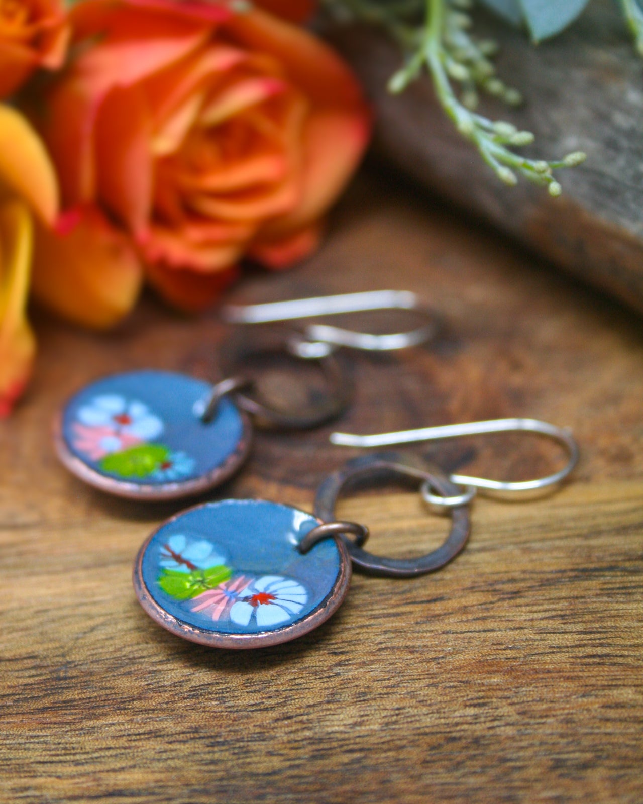 A pair of blue earrings with floral designs, resting on a wooden surface next to a bouquet of orange and yellow roses.