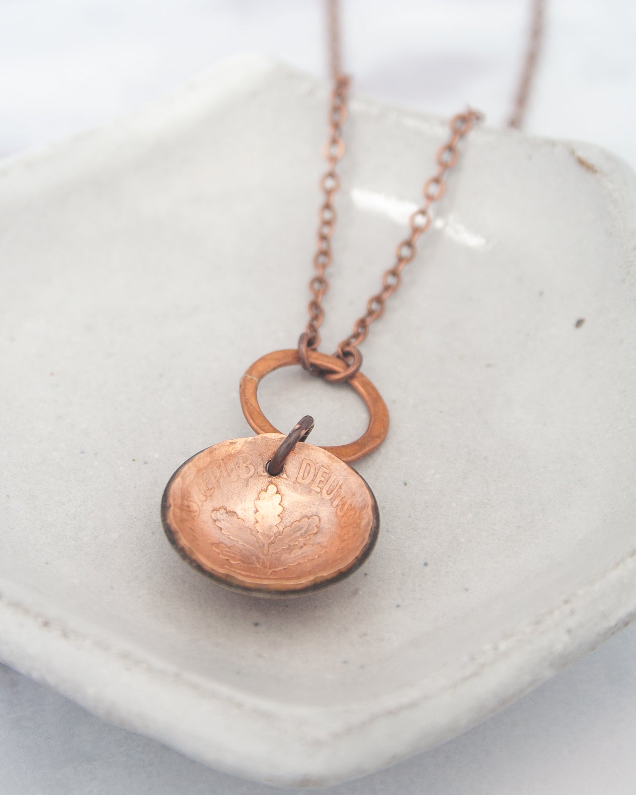 A copper-colored pendant necklace with a circular charm featuring a leaf design, resting on a white surface.