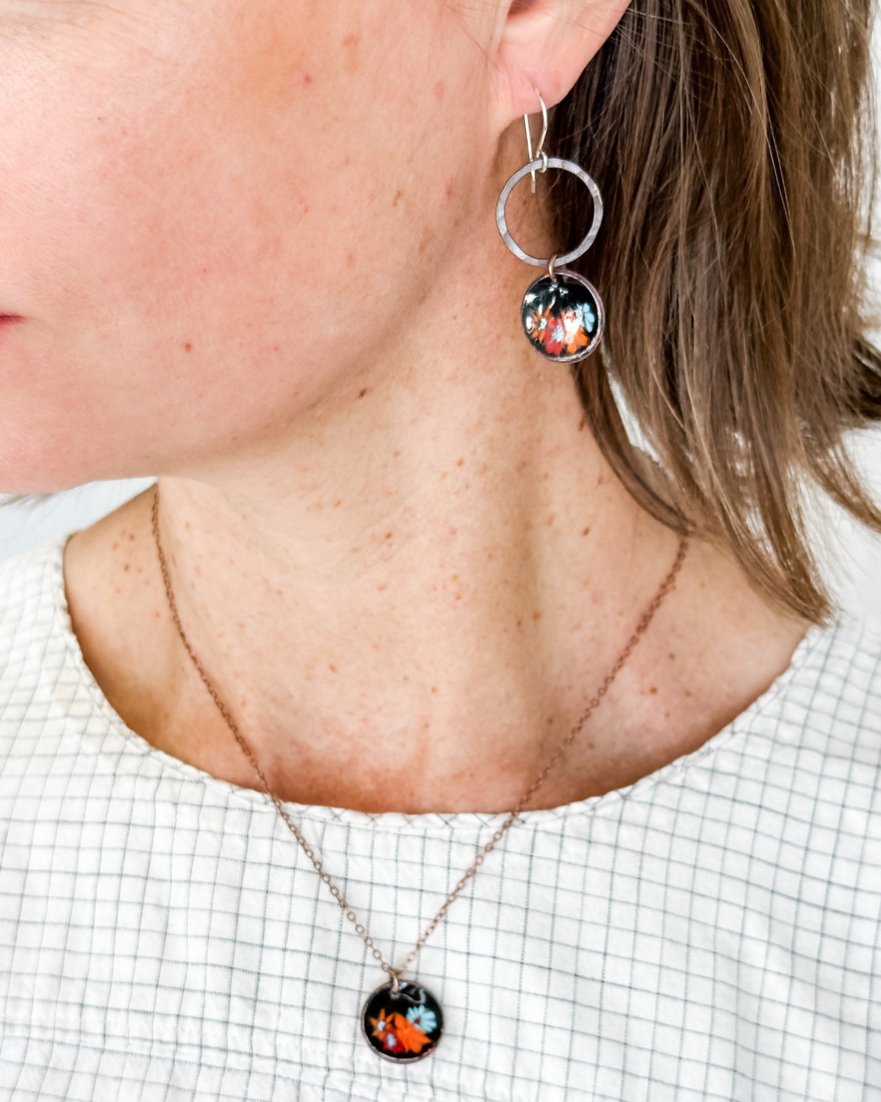 A close-up of a woman's neck and ear wearing a necklace with a pendant and earrings with a circular design.