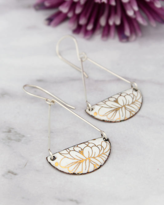 A pair of white earrings with gold floral designs, hanging from a wire and placed on a marble surface.