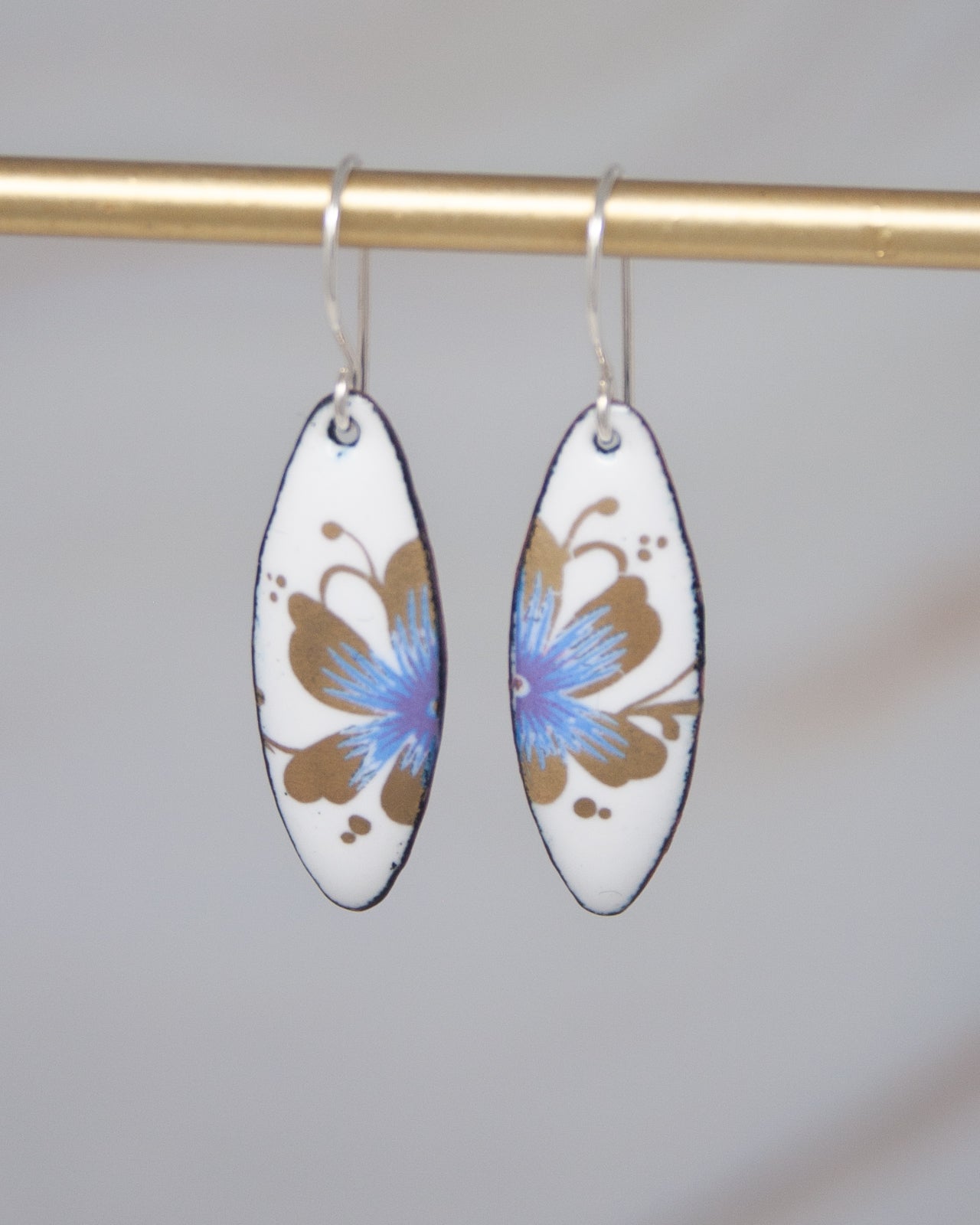 A pair of white earrings with blue and brown floral designs hanging from a gold bar.