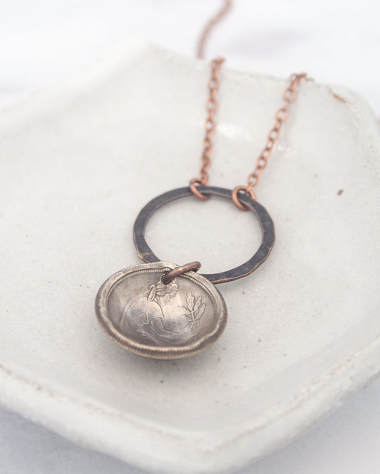 A silver-colored pendant with a circular design hangs from a chain, resting on a white surface.