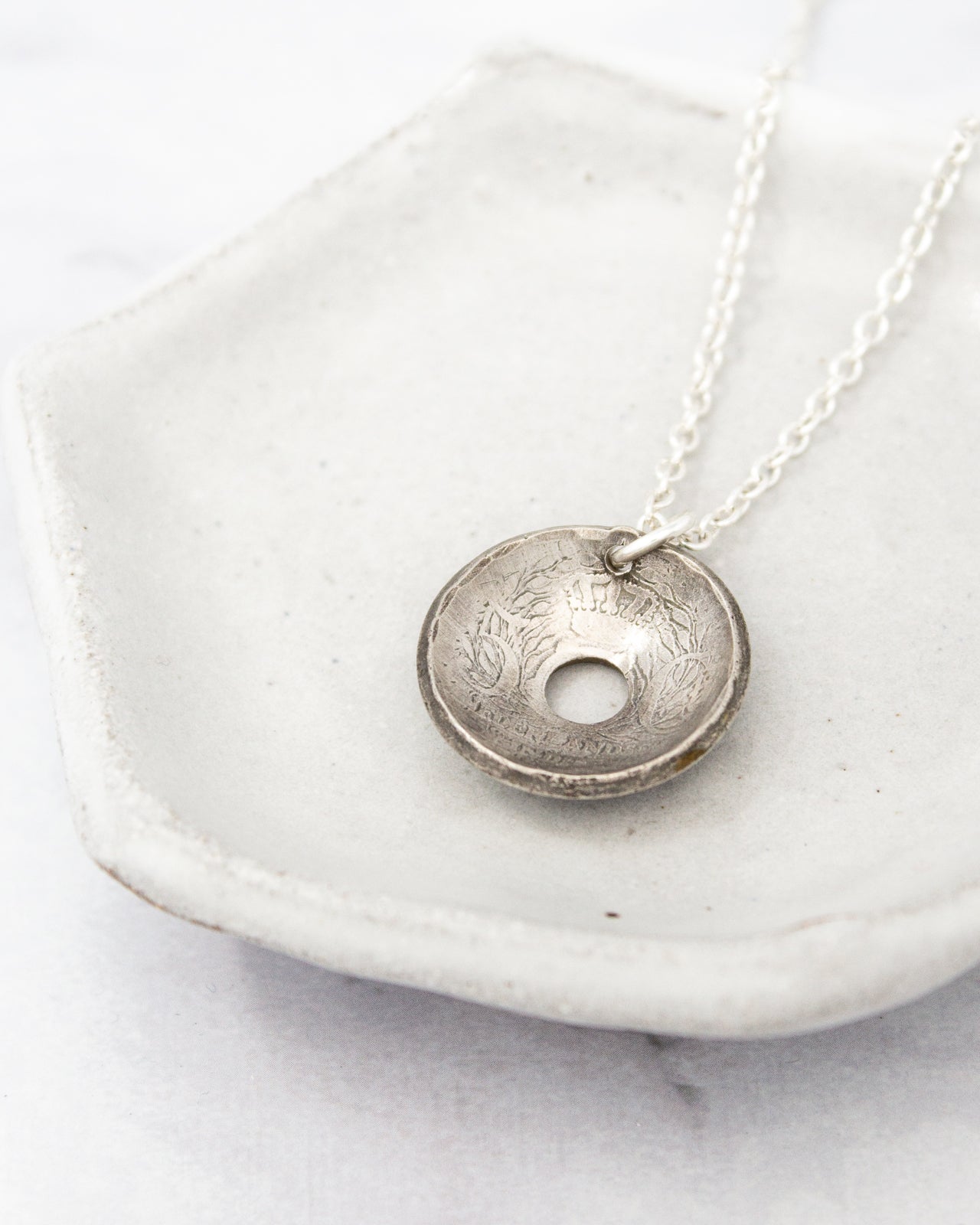 A silver pendant necklace with a circular design rests on a white ceramic dish.