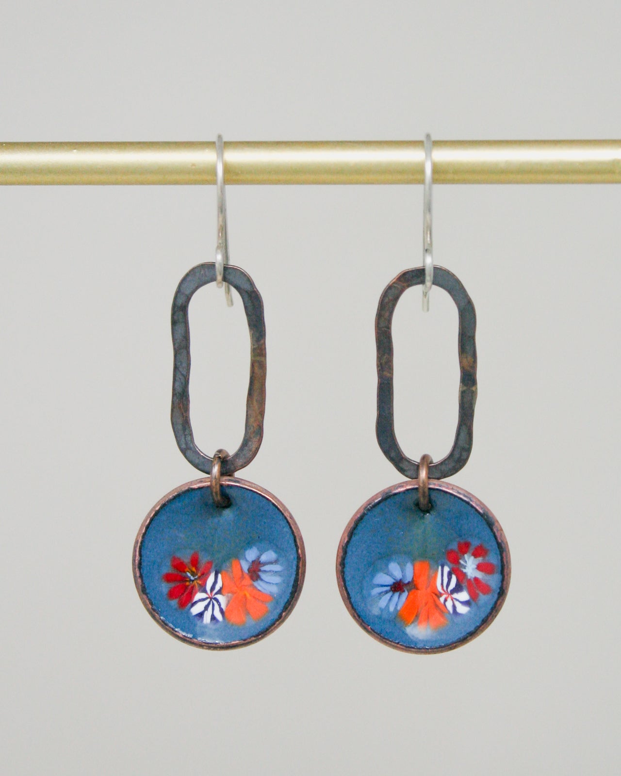 A pair of earrings with blue and orange floral designs on circular blue backgrounds, hanging from a gold rod.